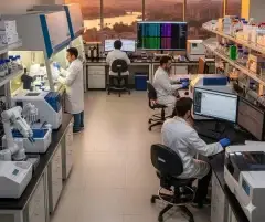 Ultra-modern and fully equipped laboratory with cutting-edge scientific instruments and equipment, including a robotic device. A large LED monitor displays critical human genetic data. One scientist works at a lab hood while others use laptops, investigating human DNA, genetics, and the DNA of healing. The image highlights the crucial role of robotic medicine in this highly important scientific field for humanity. They study how genes influence recovery, tissue repair, inflammation, and stem cell activity to understand individual differences in healing. The lab offers a stunning sunset view of the city, river, and bridge.